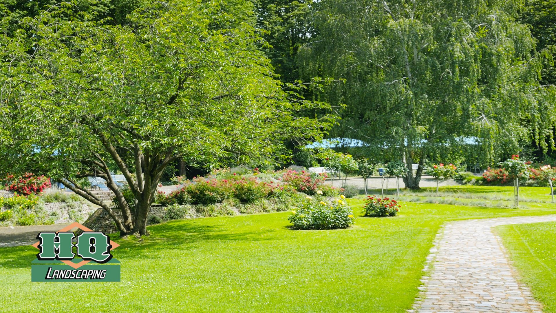 HQ lawn A beautifully landscaped park-like setting with a lush green lawn, a curved stone walkway, mature trees, and colorful flower beds, showcasing HQ Landscaping's professional lawn installation and landscaping services.